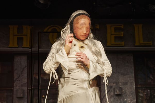 Eine Schauspielerin in weißem Kleid und hellblonder Perücke, die eine blickdurchlässige Stoffmaske trägt, auf der ein Geischt aufgedruckt ist, das leicht verschoben von dem der Schauspielerin platziert ist. Hinter der Darstellerin ist in gelben Lettern das Wort Hotel zu lesen.