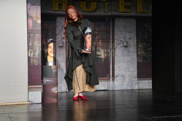 Eine Schauspielerin mit einer Maske, auf der ein Gesicht aufgedruckt ist, präsentiert eine Säule mit einem anderen Geischtsaufdruck.