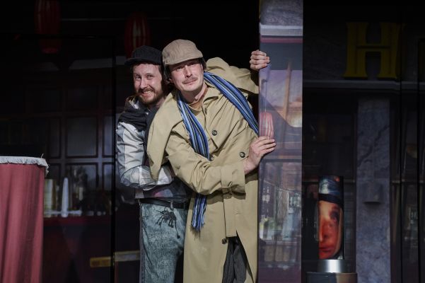 Zwei Darsteller lugen mit kritisch-skeptischen Blicken hinter einer Säule hervor.