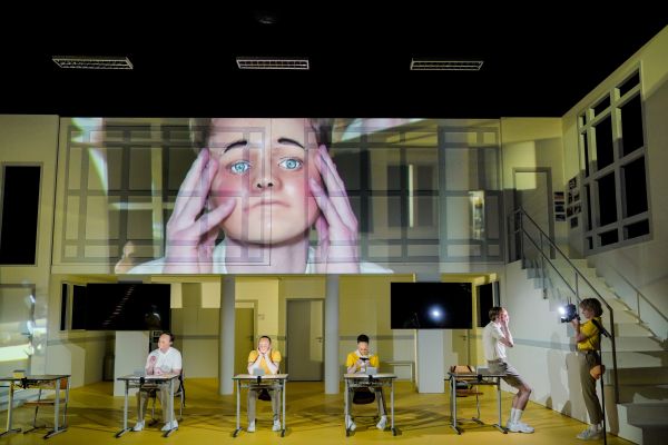 Vier Schauspielende mit Masken sitzen an Tischen in einem Klassenzimmer, eine Schauspielerin filmt das mit einer Kamera.