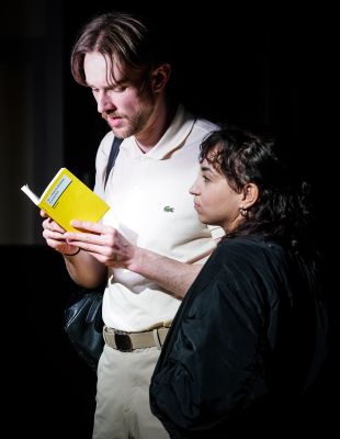 Zwei Schauspielende stehen auf der Bühne, einer liest ein gelbes Reclam Buch.