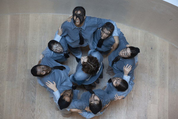 Eine Gruppe mit Feinstrumpfmasken steht im Kreis und blickt nach oben. In der Mitte steht eine Person ohne Maske.