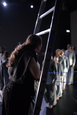 Die Regisseurin Rebecca Maria Fischer lehnt an einer Treppe und blickt das MOMO-Ensemble an.