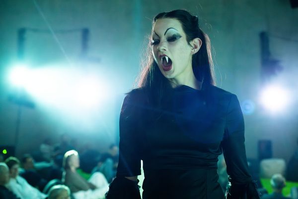 Eine Frau mit Vampirzähnen steht mit geöffnetem Mund da, im Hintergrund sieht man Menschen, die sitzen.