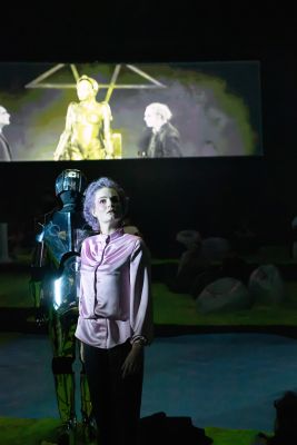 Eine auffällig gekleidete Frau und ein Roboter stehen vor einer Leinwand.