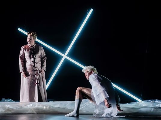 Eine Schauspielerin und ein Schauspieler vor einem leuchtenden, umgefallen Kreuz.