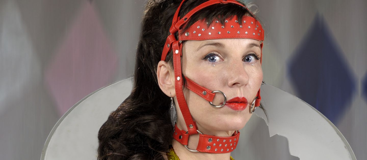 Meret Becker Meret Becker in goldenem Haltertop und auf dem Kopf ein rotes Pferdegeschirr mit Federn. Um den Hals trägt sie eine gerissene Trommel.