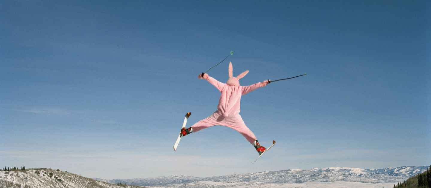 Plakatmotiv von Prana Extrem: Zu sehen ist ein Person von hinten in einem Hasenkostüm, die mit ausgestreckten Armen und Beinen mit Skiern über einem Berg springt.