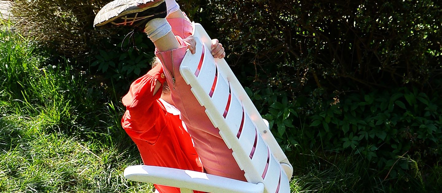 Plakatmotiv fünf minuten stille: Eine Person sitzt verkehrtherum auf einem Gartenstuhl. Die Beine sind an die Rückwand des Stuhles gelehnt und in die Höhe gestreckt. Das Umfeld besteht aus Gras und Büschen.