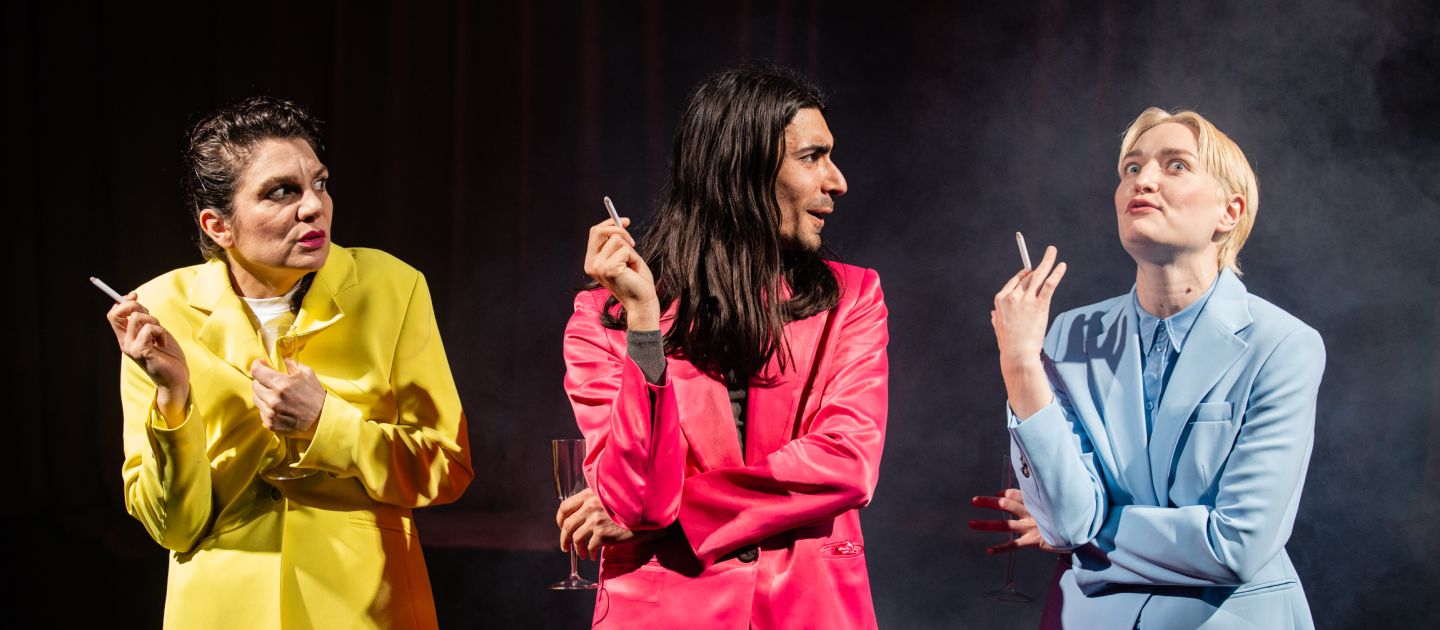Nestbeschmutzung (c) Bettina Frenzel Drei Schauspieler*innen in bunten Anzügen stehen rauchend in Reihe.