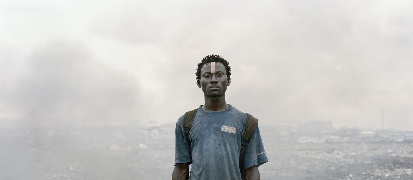 (c) Pieter Hugo, Courtesy of Stevenson, Cape Town, Johannesburg, Amsterdam Ein junger Schwarzer Mann steht mit einer Hacke in der Hand inmitten von Schutt und Rauchwolken