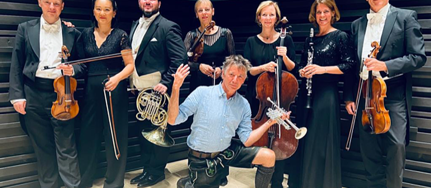 Stofferl Wells kniet auf einem Knie vor dem Ensemble, alle Personen halten ihre Instrumente in der Hand.