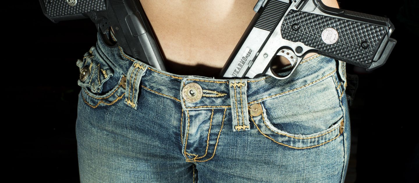 Eine Frau in Skinny Jeans und nacktem Rumpf, in dem Schritt der Hose stecken zwei Pistolen.