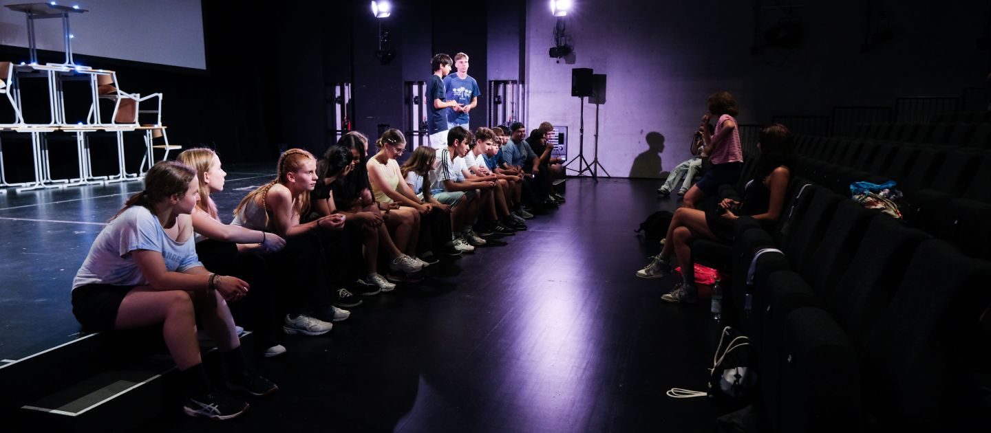 Eine Gruppe Schüler sitzt an der Bühnenkante und hört der Lehrerin zu.