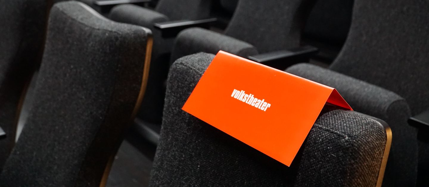 Ein roter Volkstheater-Umschlag liegt auf der Stuhlkante eines Platzes im Zuschauerraum