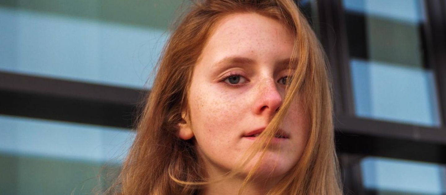 Portrait von Nathalie vor einer Fensterfront und mit Haarsträhne über den Gesicht.