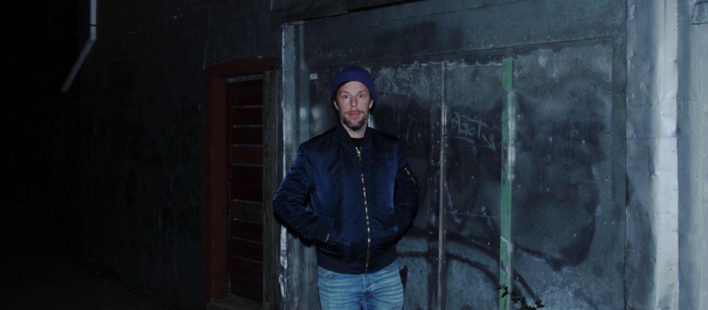 Das Bild zeigt Sebastian Pircher frontal in Jeans, dunkelblauer Jacke und Mütze vor einer dunklen Hinergrundkulisse.
