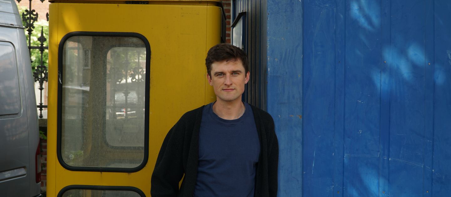 Nicholas Zöckler steht vor einer gelben Telefonzelle und einem blauen Container. 