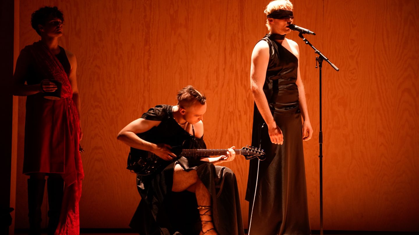 Drei Schauspieler. Einer sitzt und spielt E-Gitarre, ein anderer mit verbundenen Augen am Mikrofon.