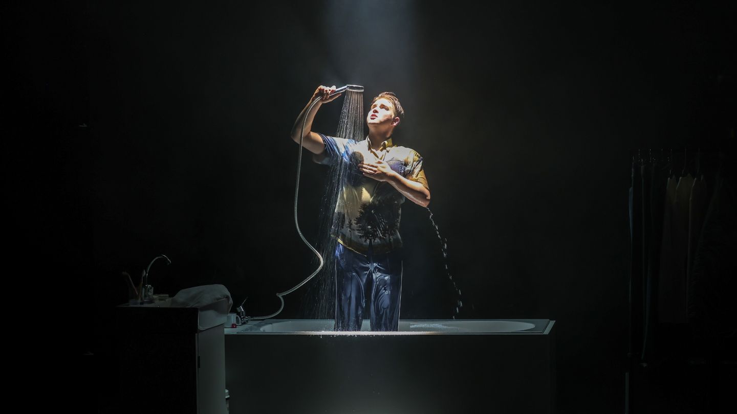 Ein Produktionsfoto von "Arbeit und Struktur". Moritz Klaus steht in einer Badewanne und duscht in seinen Klamotten.