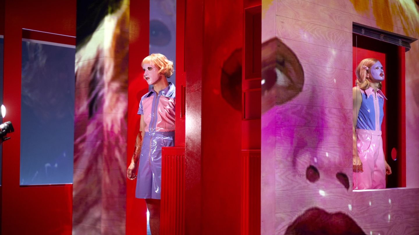 Zwei Spieler*innen mit Plastikmasken auf dem Gesicht. Ihre maskierten Gesichter werden wiederum auf die Bühnenbildwand projiziert.