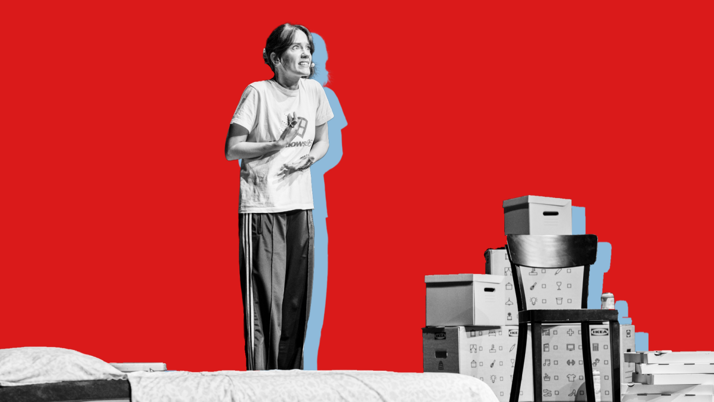 Eine Frau steht auf der Bühne, hinter ihr Umzugsboxen und ein Stuhl. 