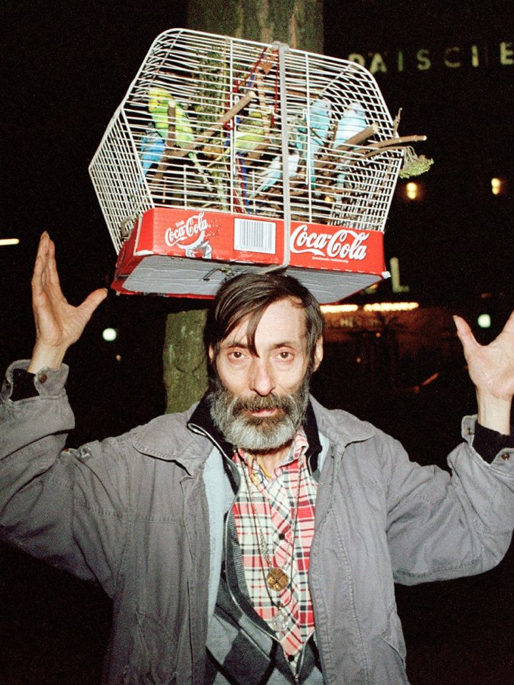 Ein Mann mit Bart. Auf seinem Kopf balanciert er einen Vogelkäfig voller Kanarienvögel.