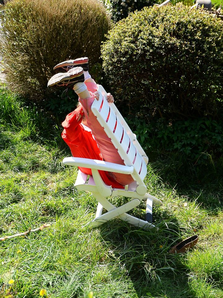 Plakatmotiv fünf minuten stille: Eine Person sitzt verkehrtherum auf einem Gartenstuhl. Die Beine sind an die Rückwand des Stuhles gelehnt und in die Höhe gestreckt. Das Umfeld besteht aus Gras und Büschen.