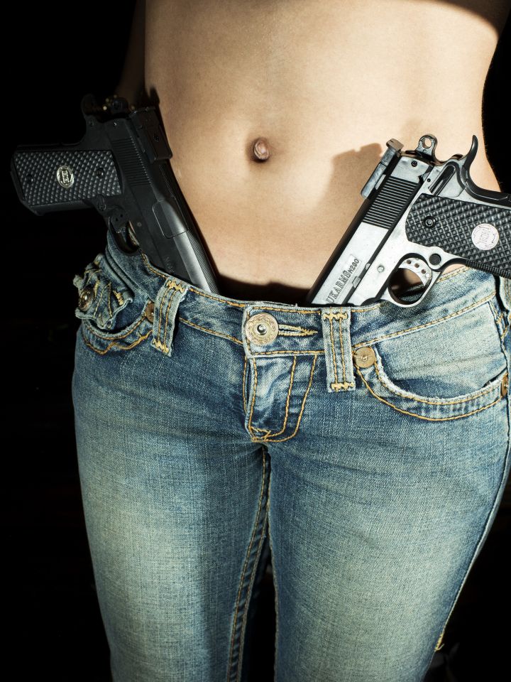 Eine Frau in Skinny Jeans und nacktem Rumpf, in dem Schritt der Hose stecken zwei Pistolen.
