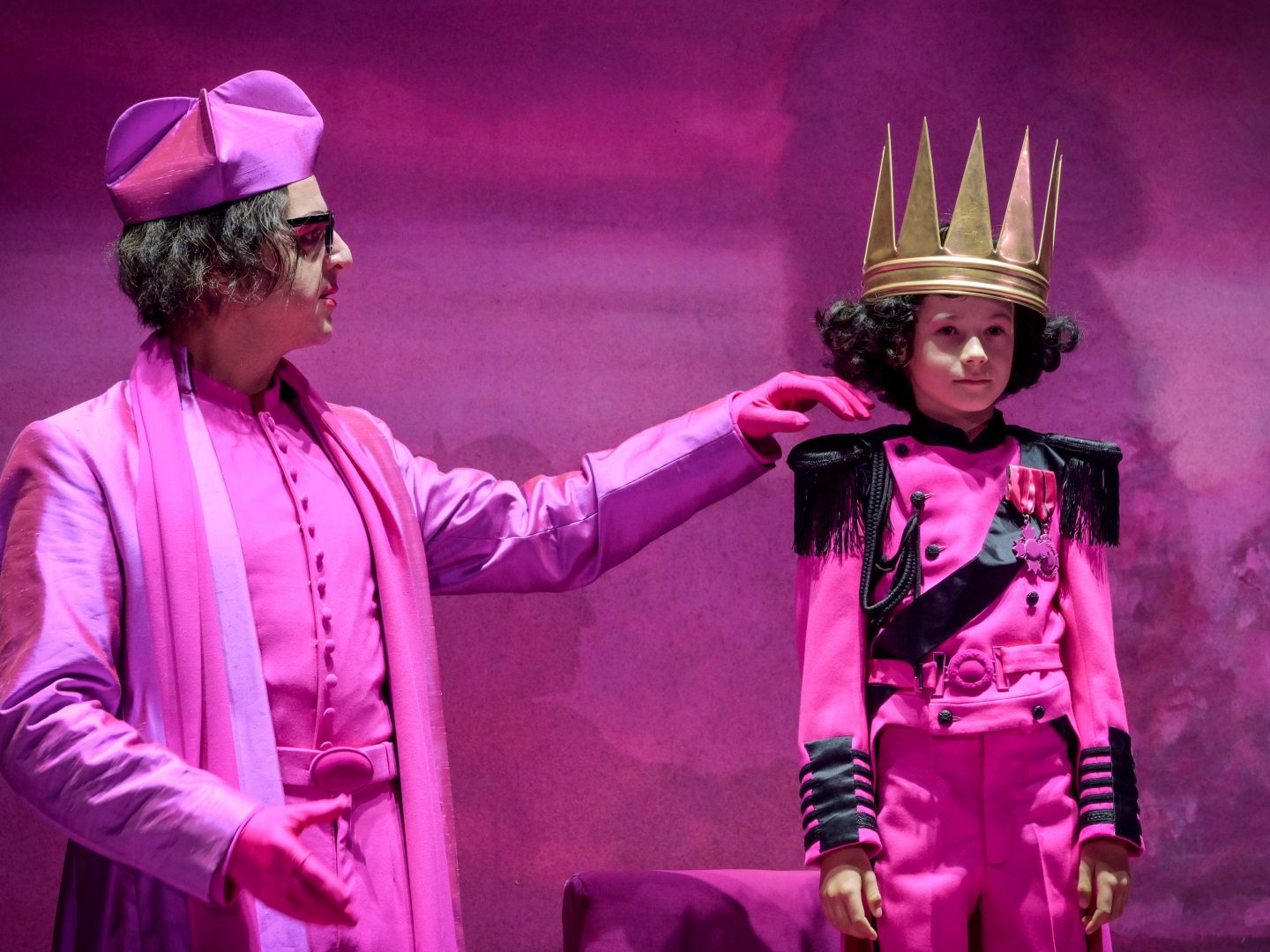 Ein in rosa gekleideter Mann streicht einem rosa gekleideten Kind mit einer Krone über die Wange.