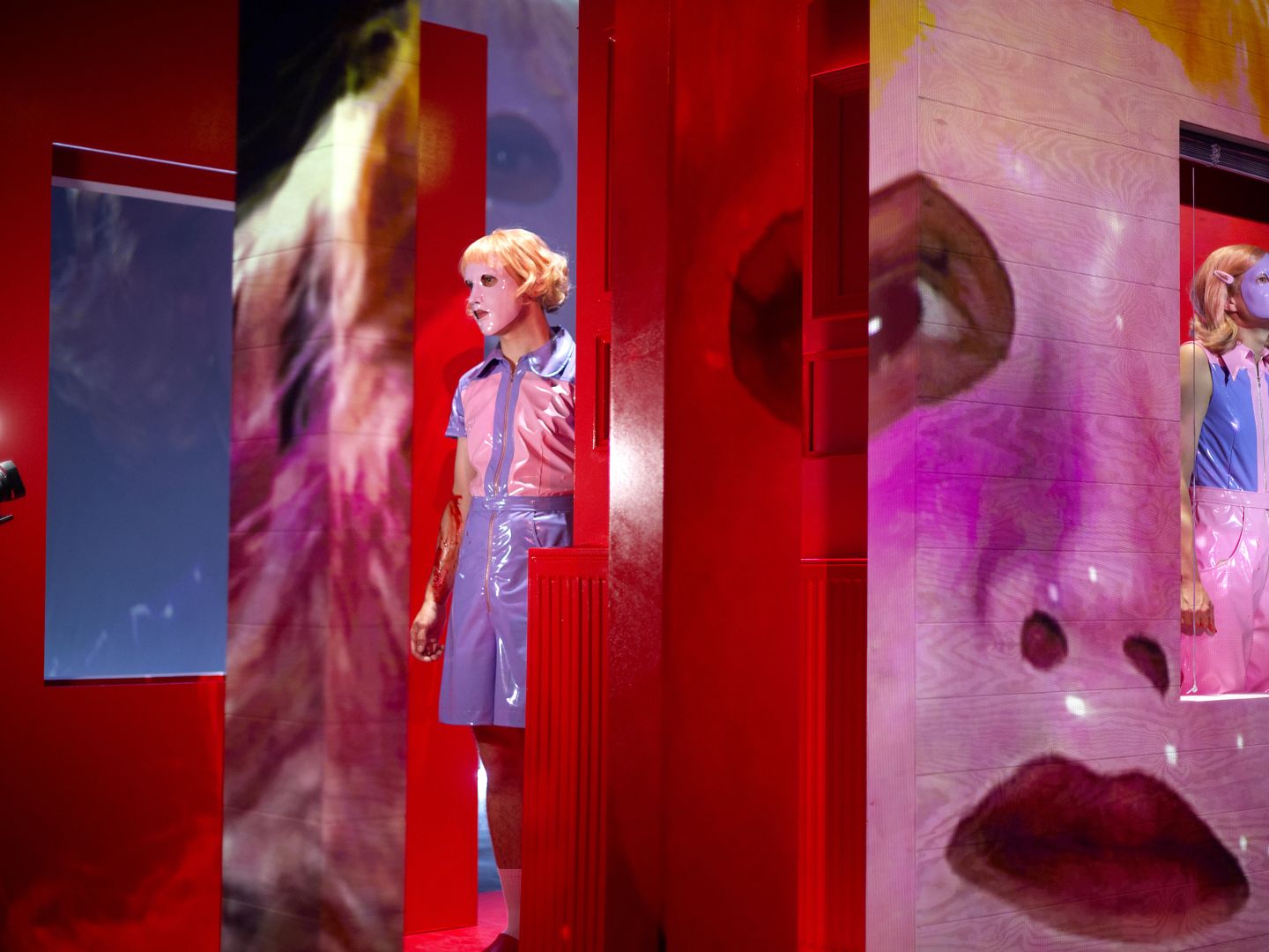 Zwei Spieler*innen mit Plastikmasken auf dem Gesicht. Ihre maskierten Gesichter werden wiederum auf die Bühnenbildwand projiziert.