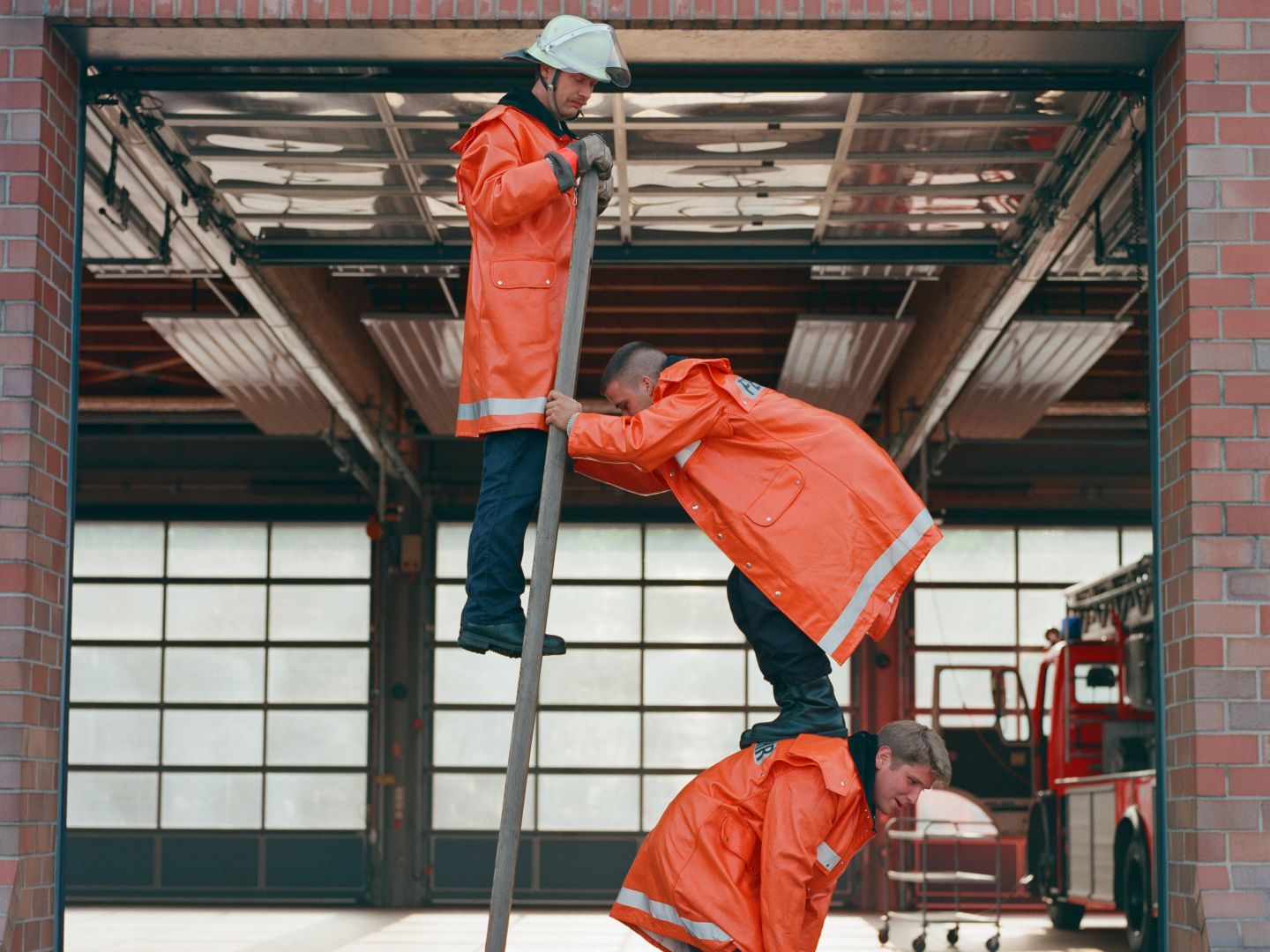 Plakatmotiv von Die Physiker. Auf dem Bild stehen drei Männer auf einem Leiter, beziehungsweise auf einander in orangenem Arbeiterkleidung .