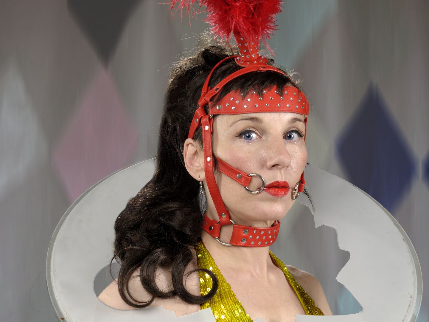 Meret Becker in goldenem Haltertop. Um den Hals trägt sie eine gerissene Trommel und auf dem Kopf ein rotes Pferdegeschirr mit Federn.