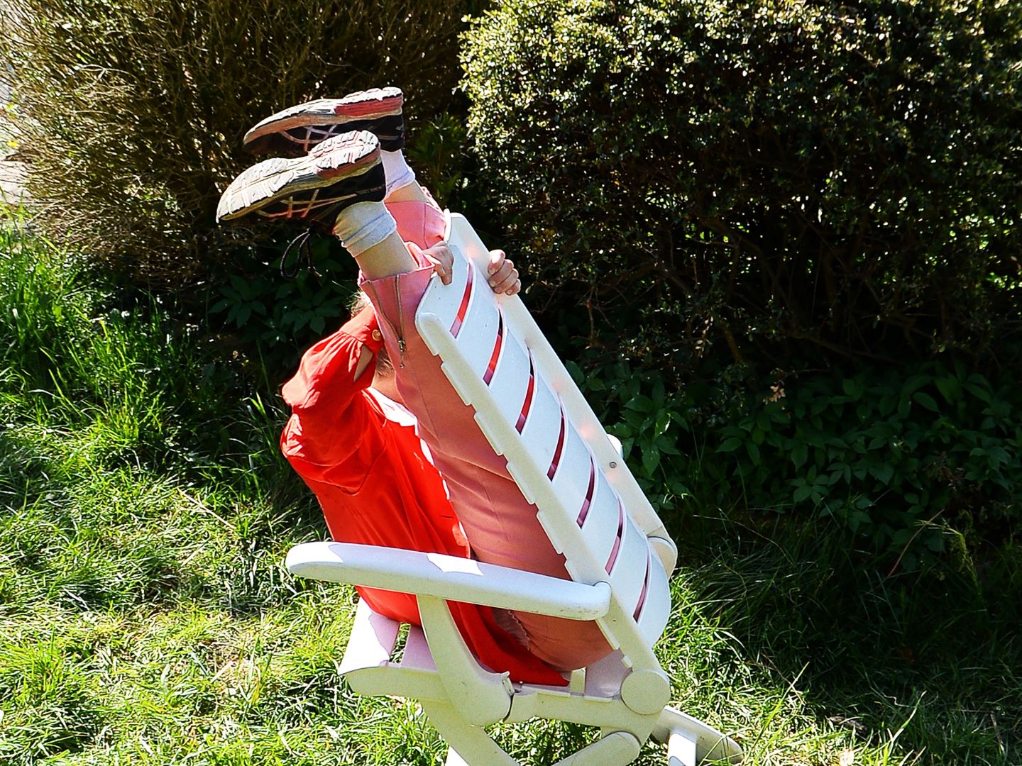 Plakatmotiv fünf minuten stille: Eine Person sitzt verkehrtherum auf einem Gartenstuhl. Die Beine sind an die Rückwand des Stuhles gelehnt und in die Höhe gestreckt. Das Umfeld besteht aus Gras und Büschen.