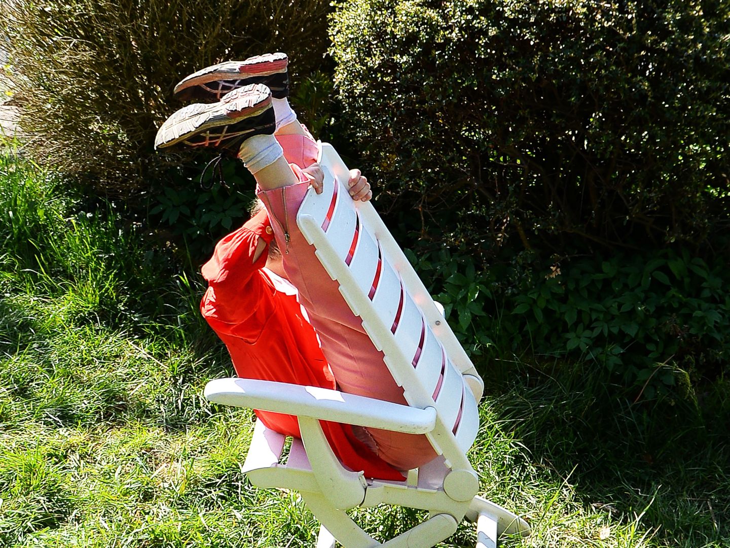 Plakatmotiv fünf minuten stille: Eine Person sitzt verkehrtherum auf einem Gartenstuhl. Die Beine sind an die Rückwand des Stuhles gelehnt und in die Höhe gestreckt. Das Umfeld besteht aus Gras und Büschen.