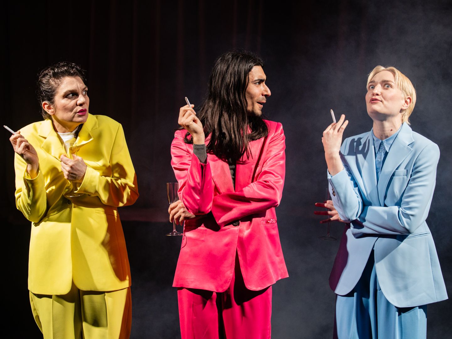 Drei Schauspieler*innen in bunten Anzügen stehen rauchend in Reihe.