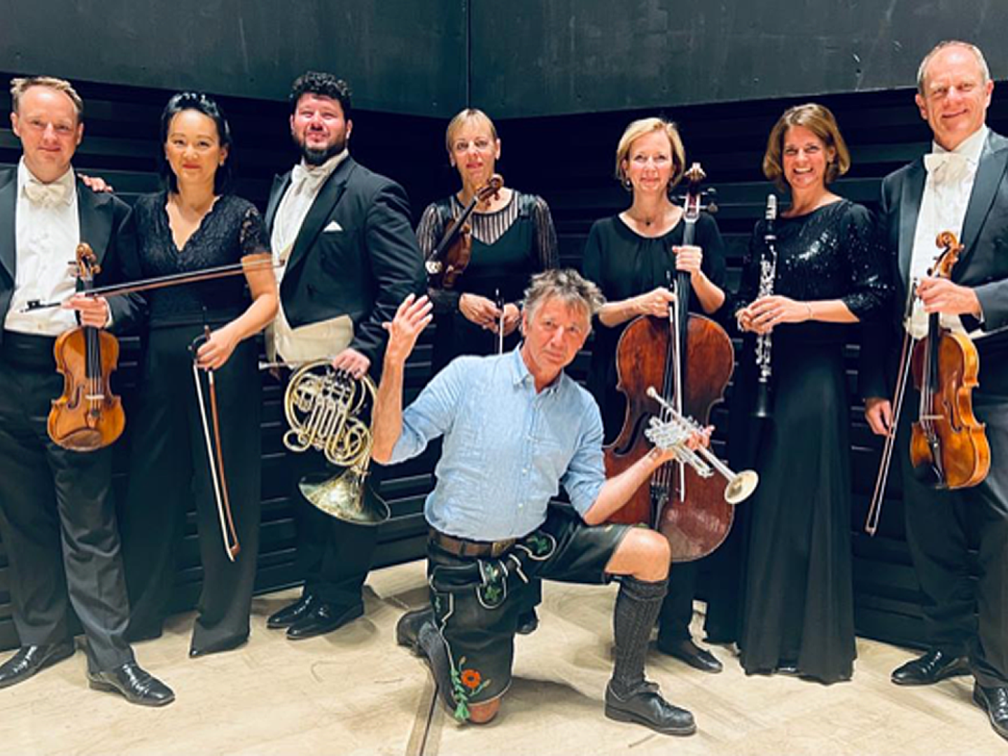 Stofferl Wells kniet auf einem Knie vor dem Ensemble, alle Personen halten ihre Instrumente in der Hand.