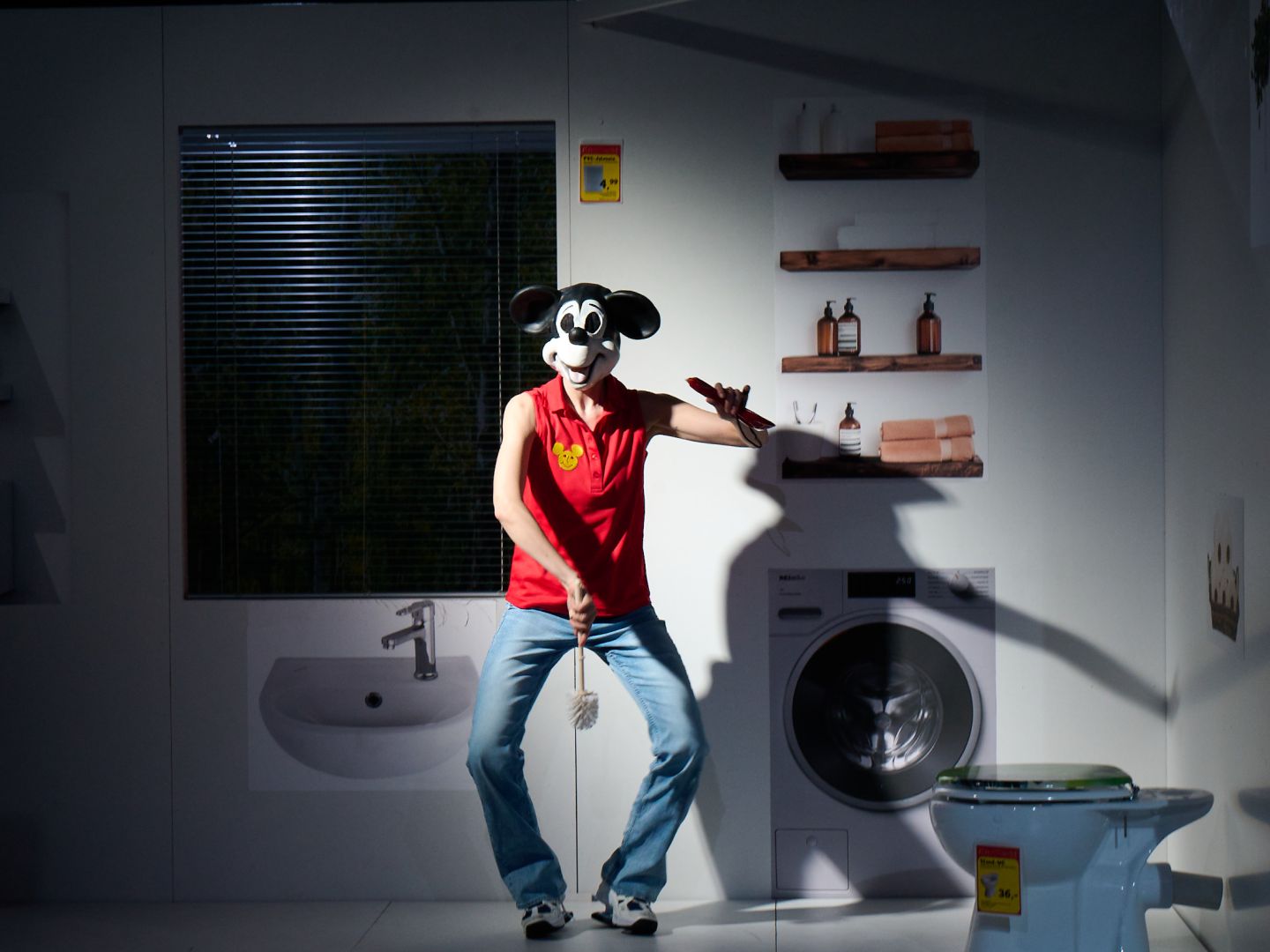 Luise Hart in Jeans und rotem Tanktop, bedruckt mit einem kleinen gelben Mickey Mouse Kopf, steht tänzerisch mit O-Beinen und hält eine Klobürste mittig vor sich. Auf dem Kopf trägt sie einen Mickey Mouse Kopf.