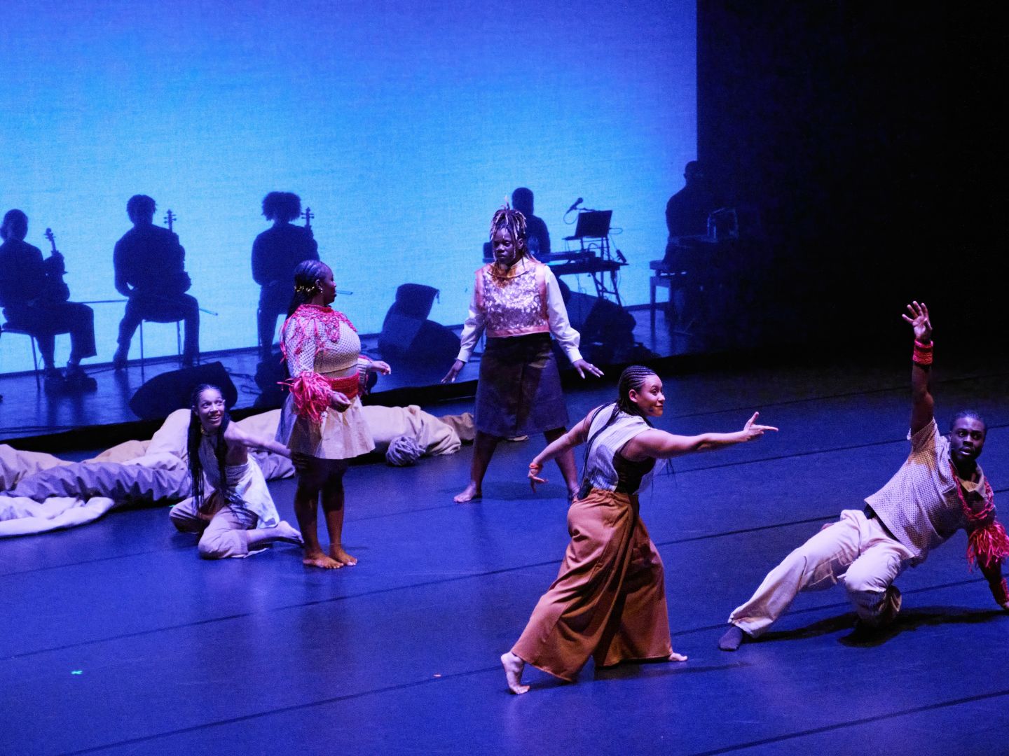 Ensemblemitglieder in tänzerischen Bewegungen. Hinter ihnen sind Schatten von Musiker:innen hinter einem transparenten Vorhang sichtbar.