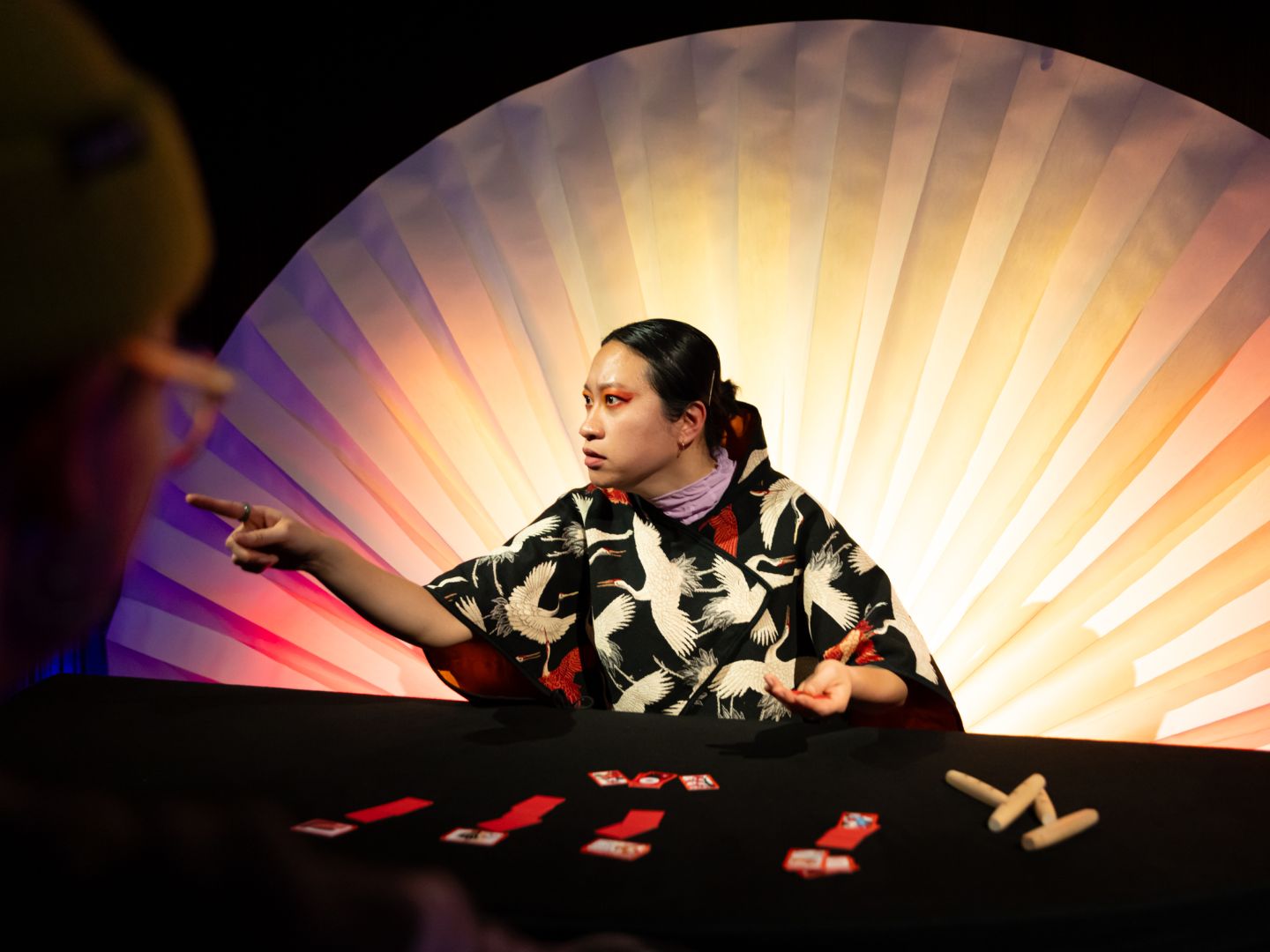 Olivia Hyunsin Kim sitzt in einem mit Pelikanen bedruckten Kimono und auffälligem orangen Augenmakeup vor einem großen weißen Fächer an einem Tisch und deutet auf eine Person außerhalb des Bildes. Vor ihr liegen etliche rote Karten mit Motiven.