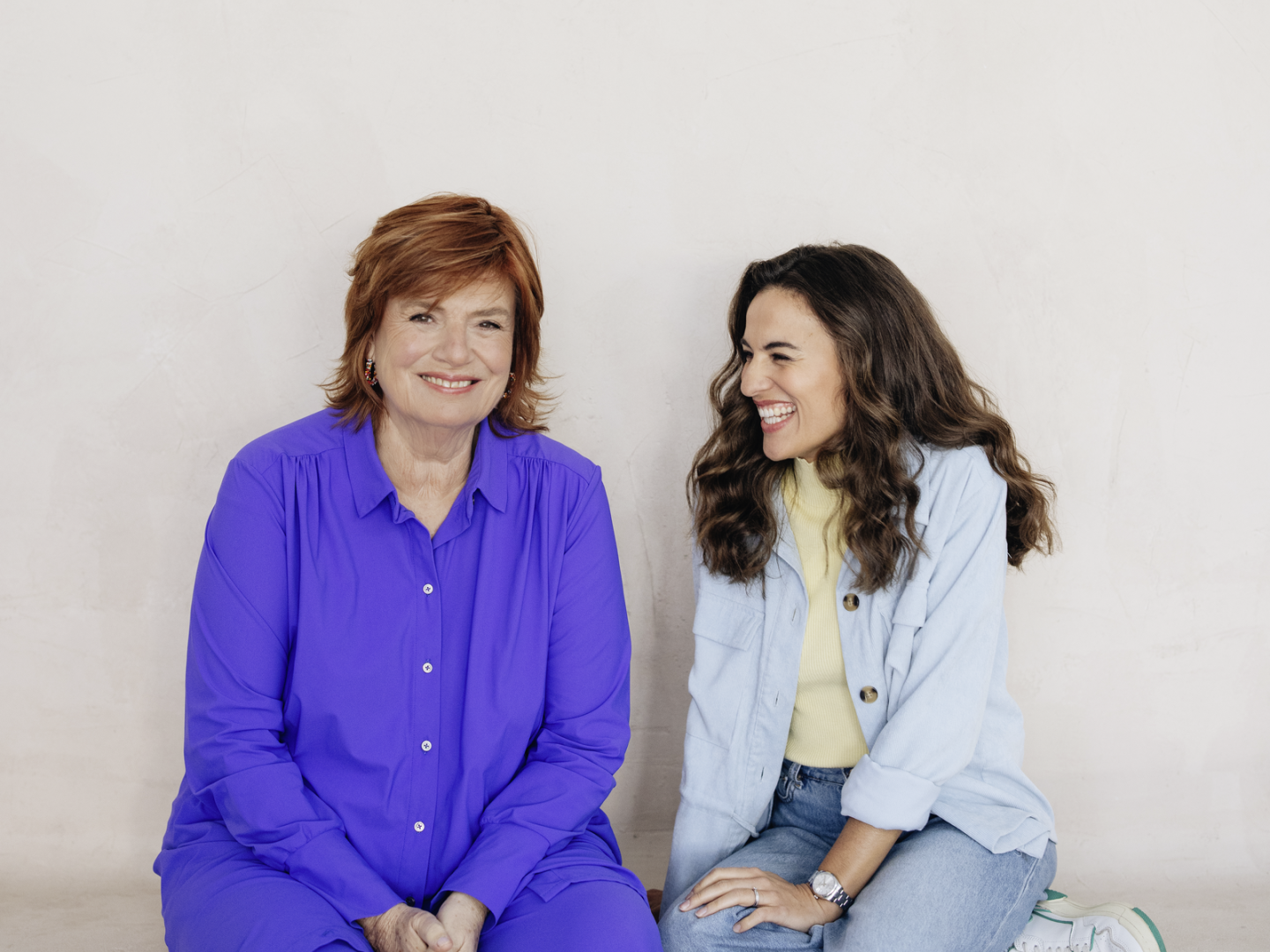 Mona Ameziane &amp; Christine Westermann lachend vor einer grauen Wand sitzend.