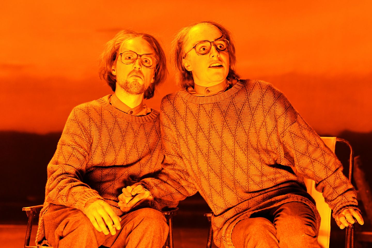Das Foto zeigt zwei Männer in identischen Pullovern, die nebeneinander auf Stühle sitzen und sich an den Händen halten.