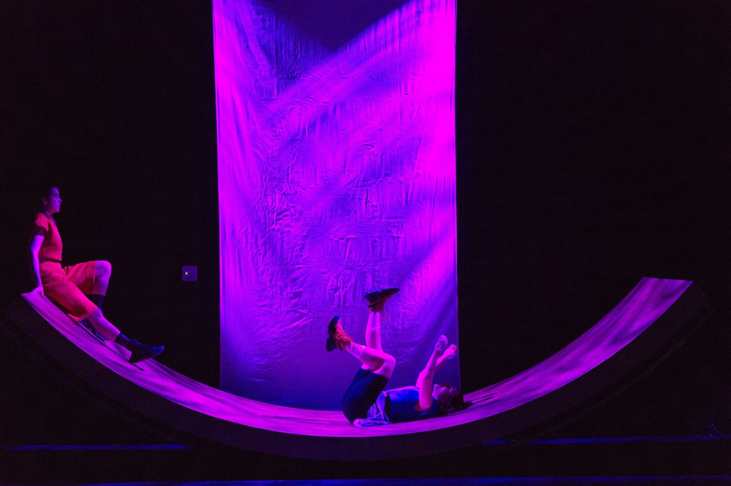 Das Bild zeigt zwei Personen auf einer großen Wippe. Eine sitzt an einer oberen Kanten und blickt zu der anderen Person, die strampelnd in der Mitte liegt.