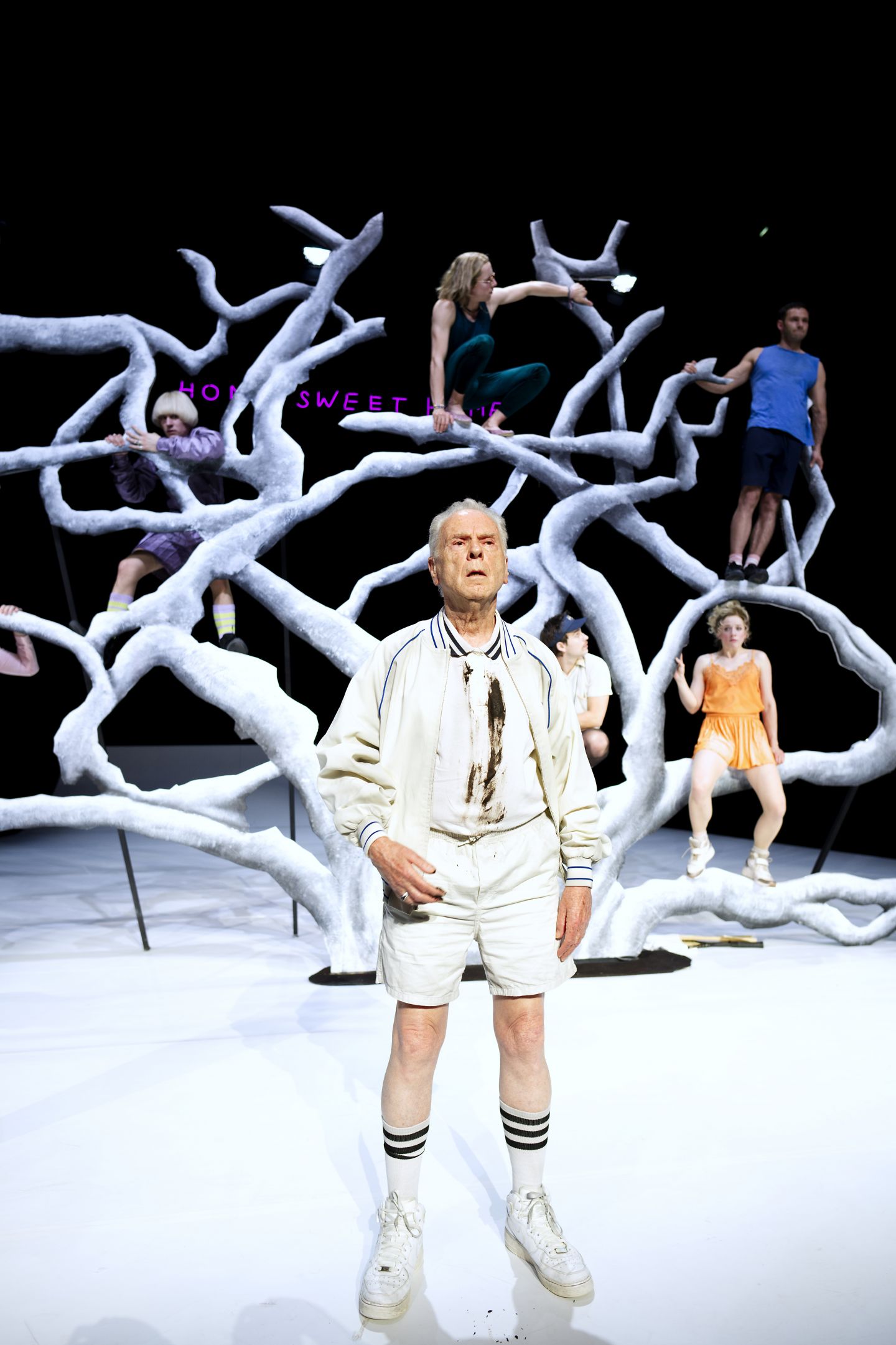 Hans Dieter Trayer in seiner Rolle steht ganz in weiß vorne auf der Bühne. Im Hintergrund ist das Ensemble sitzend auf dem weißen Baum zu sehen.