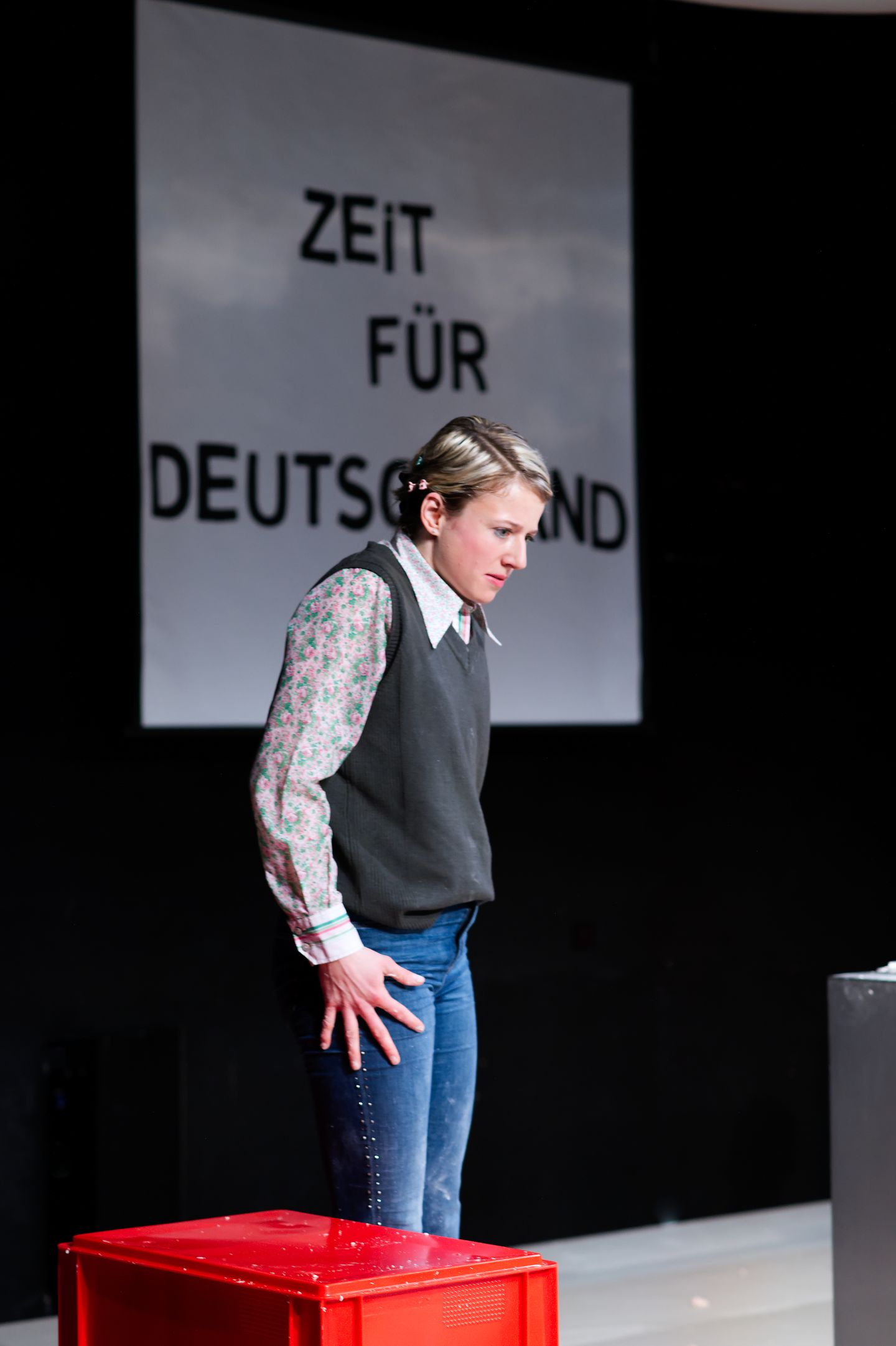 Die Rolle von Henriette Nagel steht in Jeans und Bluse in der Mitte der Bühne. Ihr Schultern sind hochgezogen, ihre Hände ausgespreizt seitlich am Körper und sie blickt auf dem Boden. Im Hintergrund ist ein Plakat mit der Aufschrift "Zeit für Deutschland" zu sehen.
