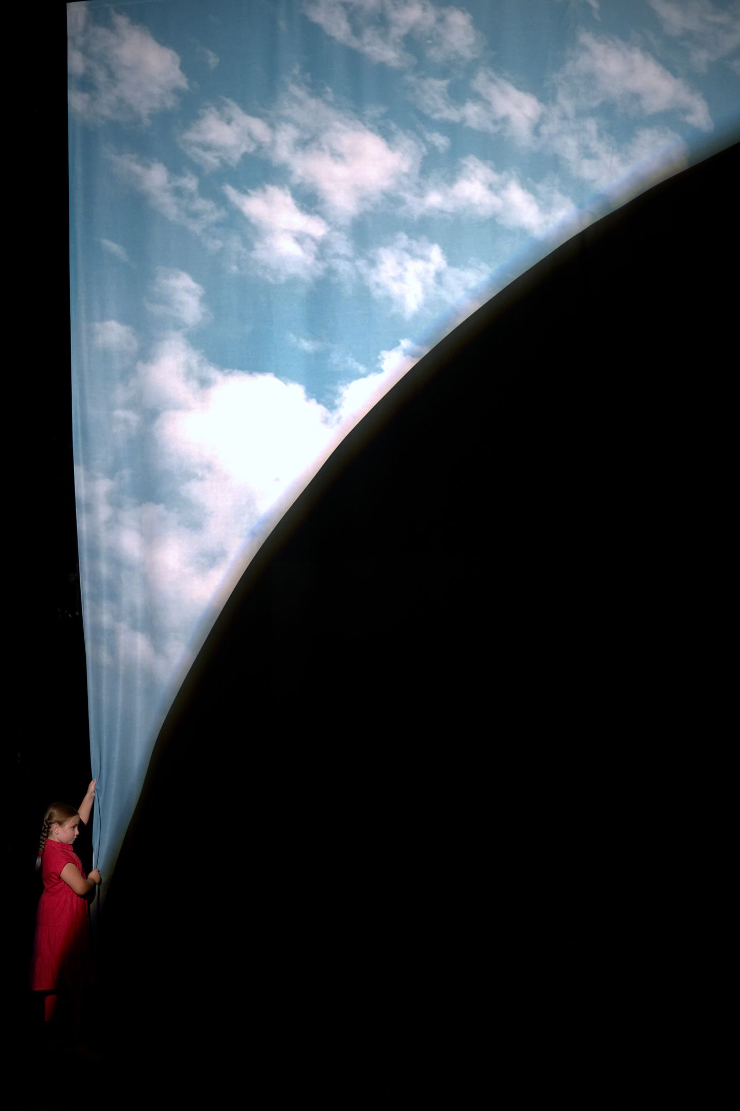 Käthe Haddad zieht einen großen Vorhang zur Seite auf dem ein bewölkter Himmel zu sehen ist.