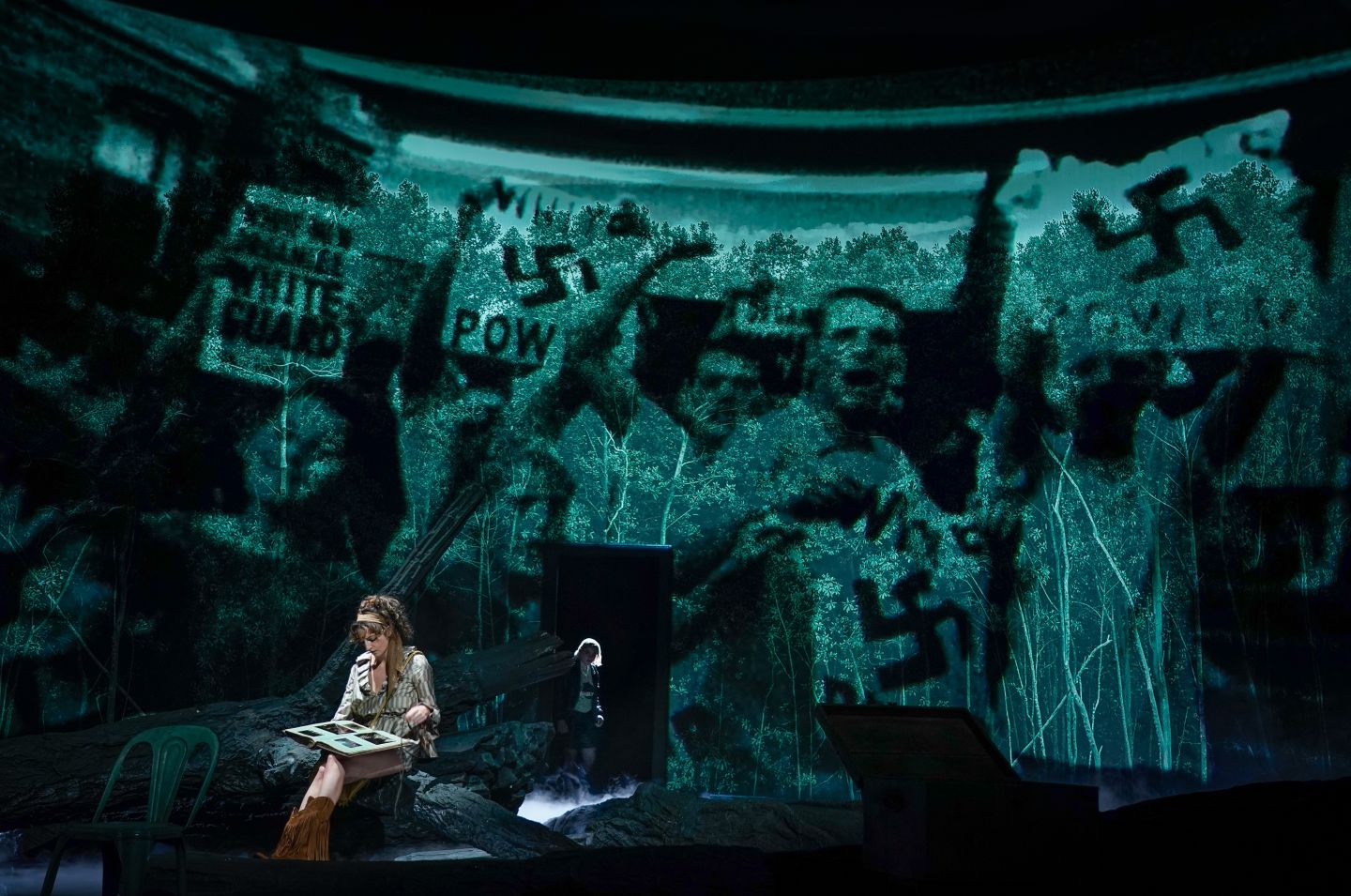 Marlene Markt betrachtet, auf einem Baumstamm sitzend, ein Fotoalbum. Im Hintergrund wird eine Aufnahme von einer White Power Demonstration projiziert. Lasse Stadelmann ist durch einen Eingang zur Bühne ebenfalls im Hintergrund zu sehen.