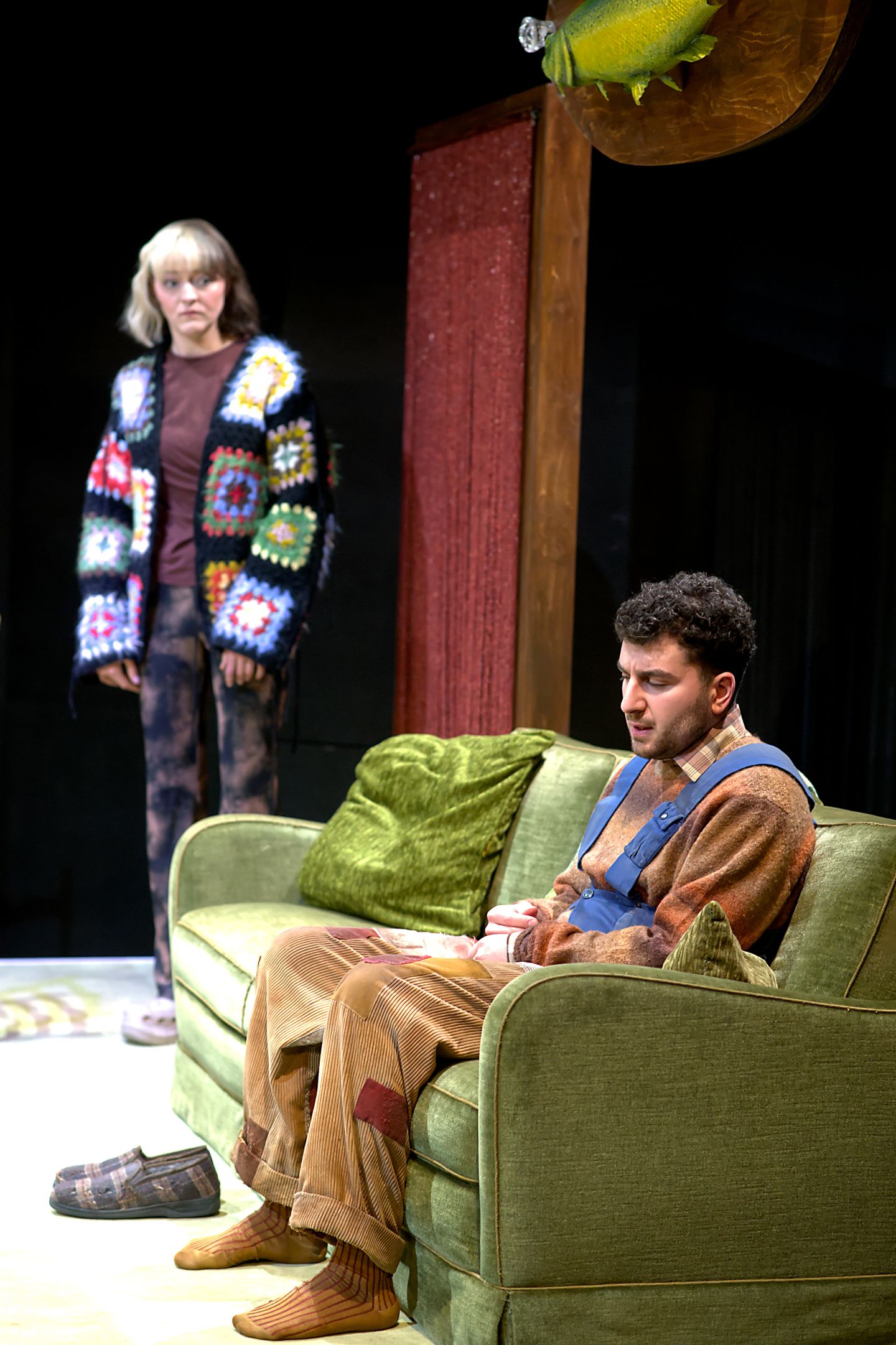 Eine Schauspielerin steht, ein Schauspieler sitzt auf der Couch.