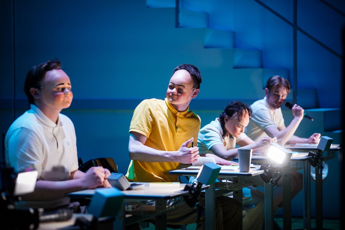 Vier Schauspieler*innen mit kindlichen Masken sitzen an Schulbänken.