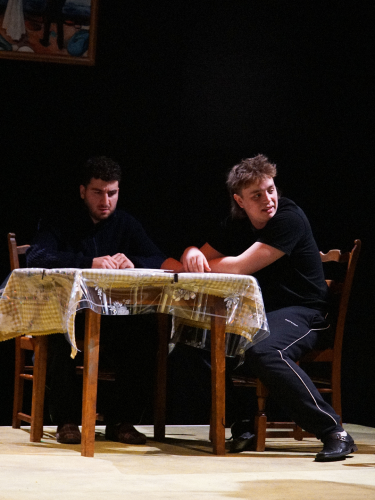 Eine Theaterprobe: Zwei Schauspieler sitzen an einem Tisch blicken skeptisch zur Seite.